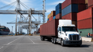 Container truck near port cranes and stacked shipping containers for drayage services.